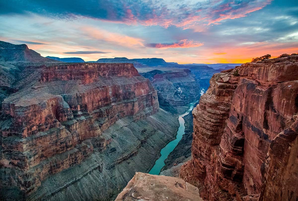 Looking down past sheer cliff walls 3,000 vertical feet (915 m) to a river flowing below. Sunset clouds are tinged with brilliant pink.