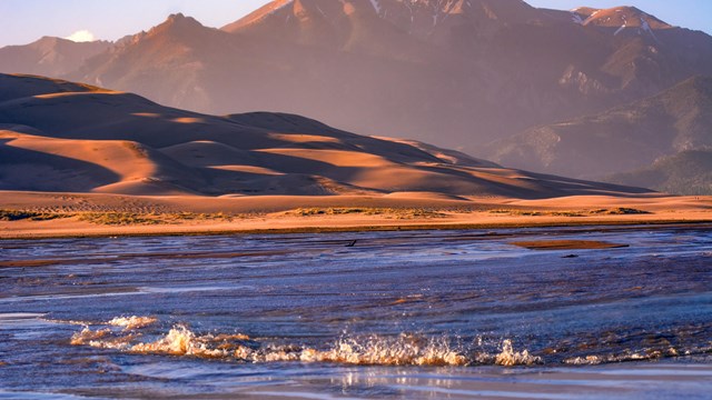 A shallow creek with a wave in front of dunes and a mountain