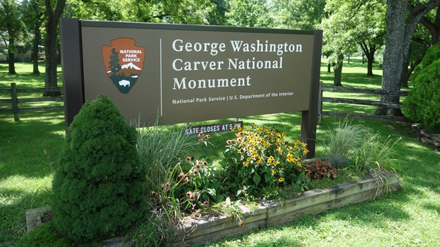 Park entrance sign to the with a National Park Service arrowhead & flowers.