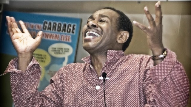 A man wearing a red shirt and holding his hands in the air with a lapel mic. 