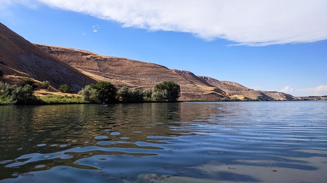 Photo of rocky, golden bluffs sloping down toward the Snake River, with a small kayaker on the water