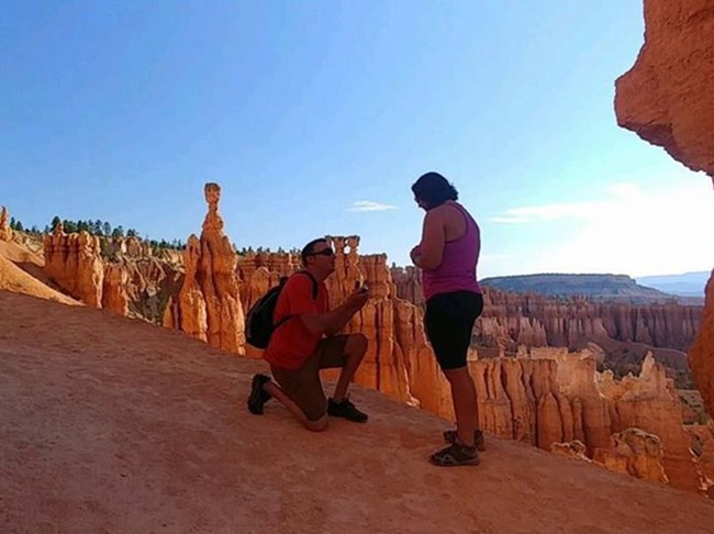 A man on one knee holds a box with a ring in it and looks up at a woman