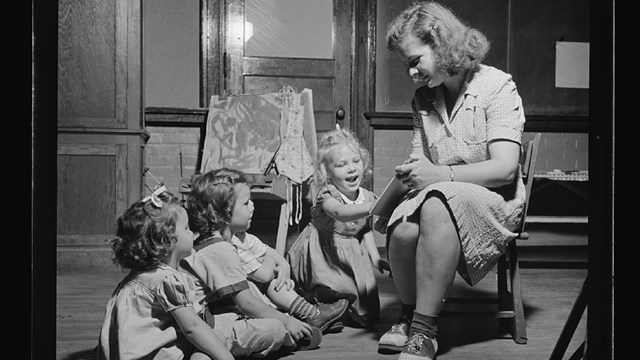 Three white girls sit on the floor looking at a book held by a white woman sitting in a chair