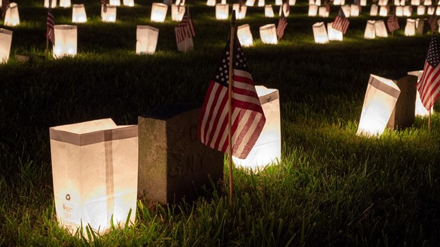 Luminaries surrounding rows and rows of soldier graves decorated with small flags at night 