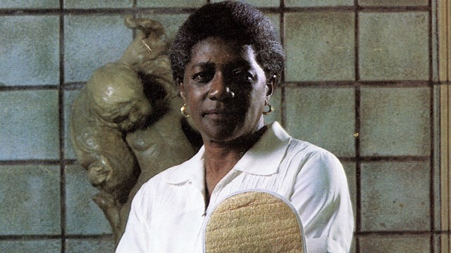African American woman wearing white shirt and holding towel and scrub stands indoors.
