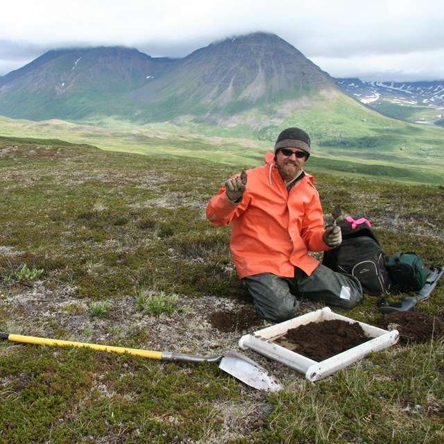 A man in orange jacket kneels in front of a screen with dirt in it, with cloud capped mountain peaks