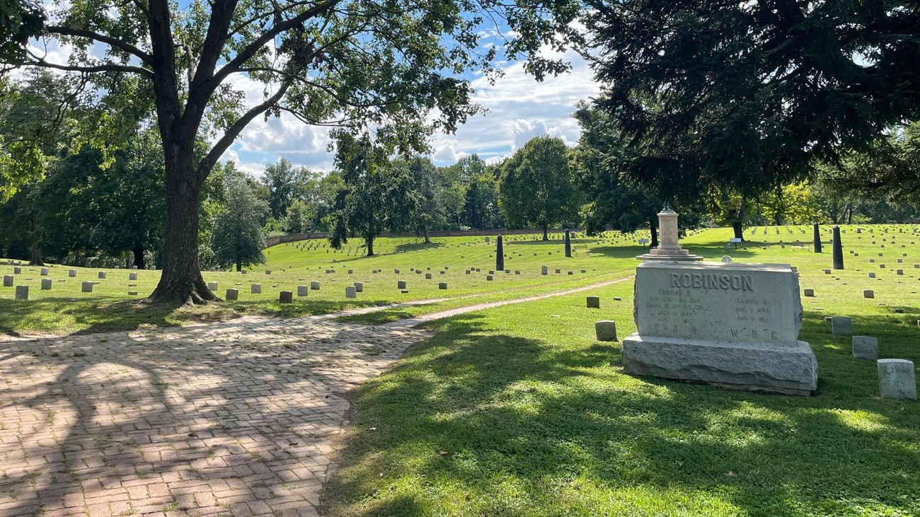 Dappled shade on a brick path beside a leafy tree, beside rows of uniform headstones