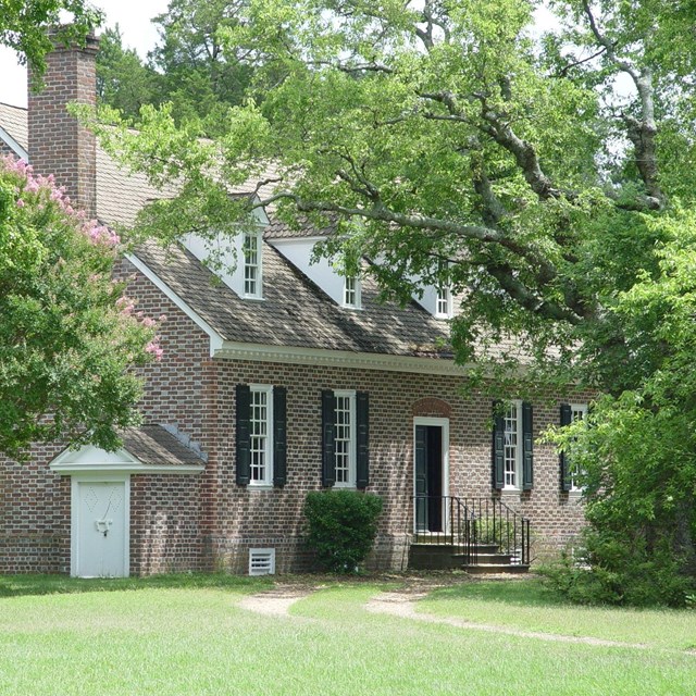 A large brick building, with a sloped roof and dormers. It is surrounded by green trees and grass.