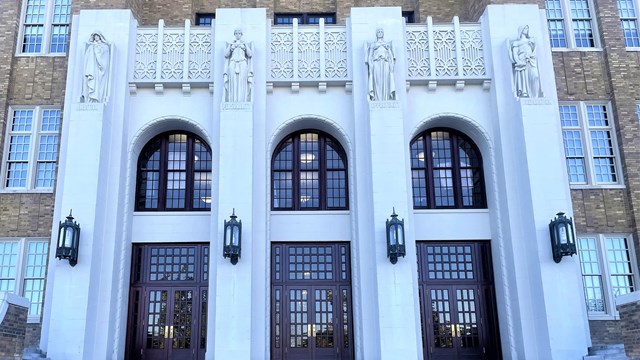 The main entrance is flanked by arched windows, lantern fixtures, & statues above 3 sets of doors.