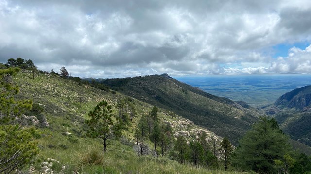 A tall peak is seen at the end of a long ridge with clouds in the background