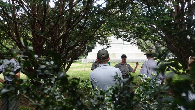 A view through an opening in a tall shurb reveals NPS staff, with a monument partly visible beyond.