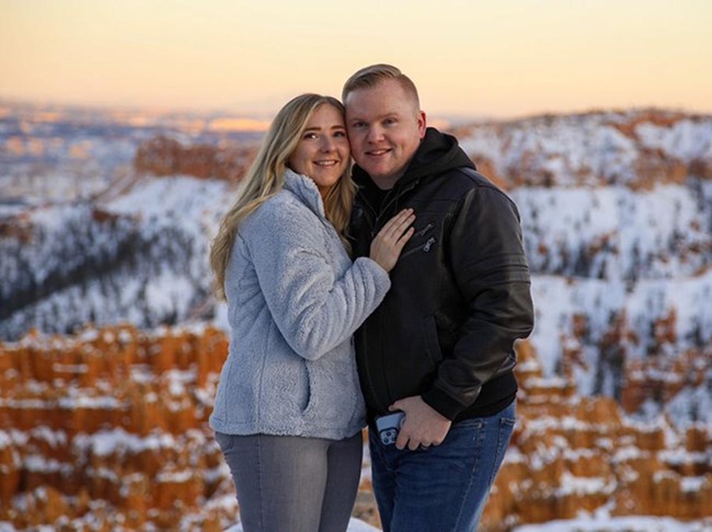 A man and woman face the camera with a blurred background of red rocks behind them