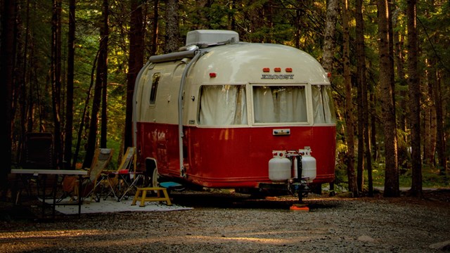 Photo of a white and red vintage trailer under trees at a campsite. 