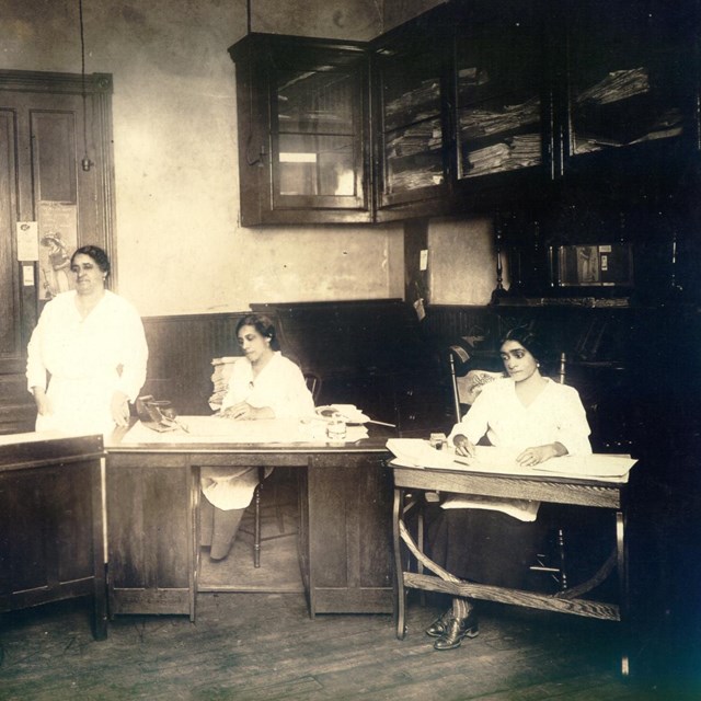 B&W Photo; 3 women work at desks, maggie walker stands