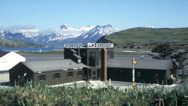 a squat, L-shaped building sits at the base of a coastal range with snowy mountains.