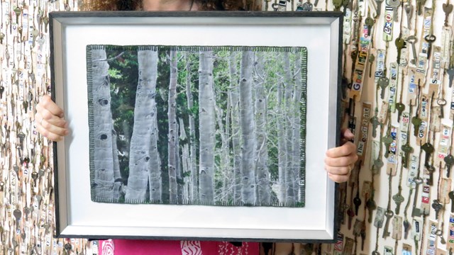 A color photo of a woman with long curly hair holding a painted quilt depicting green/white aspens