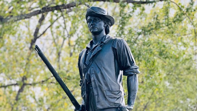 A bronze statue of a minute man sits atop a granite base. The man is walking and carrying a musket