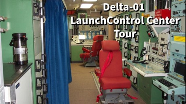 A short narrow room with green and gray computer racks and two red chairs