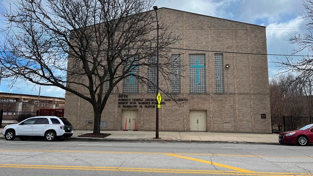 A multi-story brick church stands along the side of a street. A tree stands in front. 