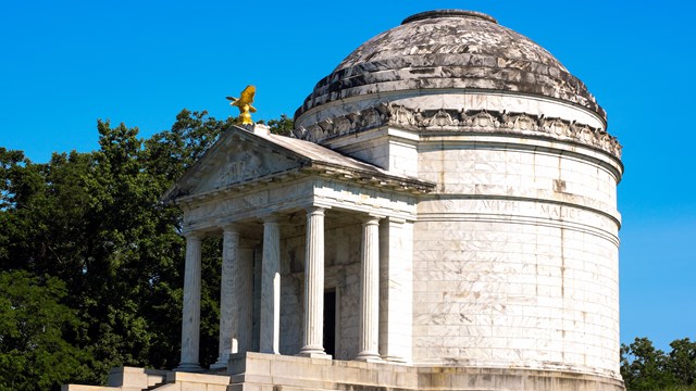 A large white marble structure with a dome roof sits atop a small grassy hill.