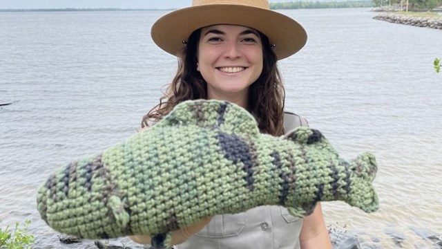 A park ranger stands in uniform holding out a walleye crocheted out of yarn.