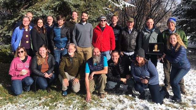 Photo of a couple dozen smiling people standing in grass in front of tree