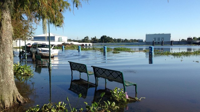 Two green benches, water flows beneath, vehicle with boat ready to launch into a flooded river.