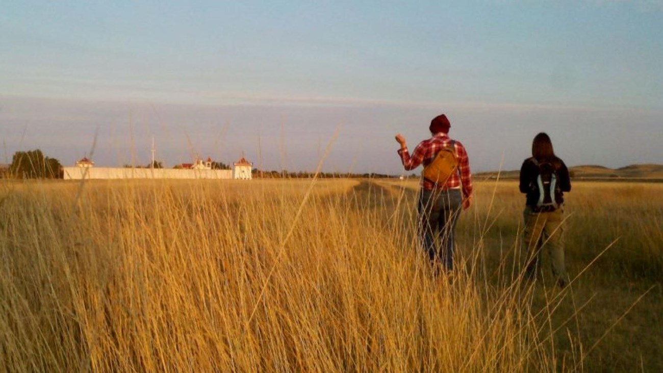 Two people walk in a field towards a white walled Fort. 