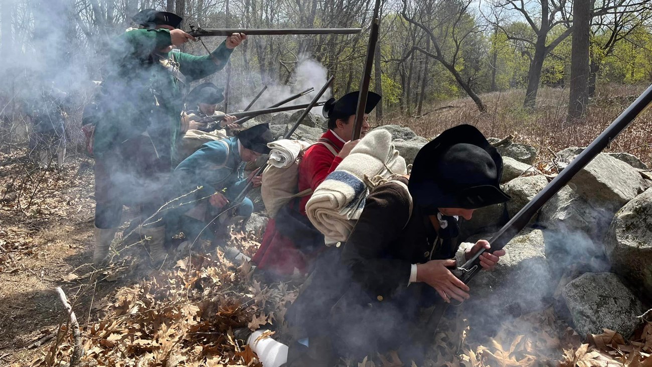 Militia soldiers crouch behind a stone wall loading and firing muskets while surrounded by smoke.