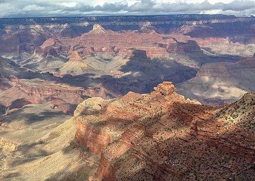 on an overcast day with dramatic shadows, a view down a ridgeline of vermilion colored stratified rock to a lone promontory. In the distance peaks and cliffs are seen rising from the floor of a mile-deep canyon.