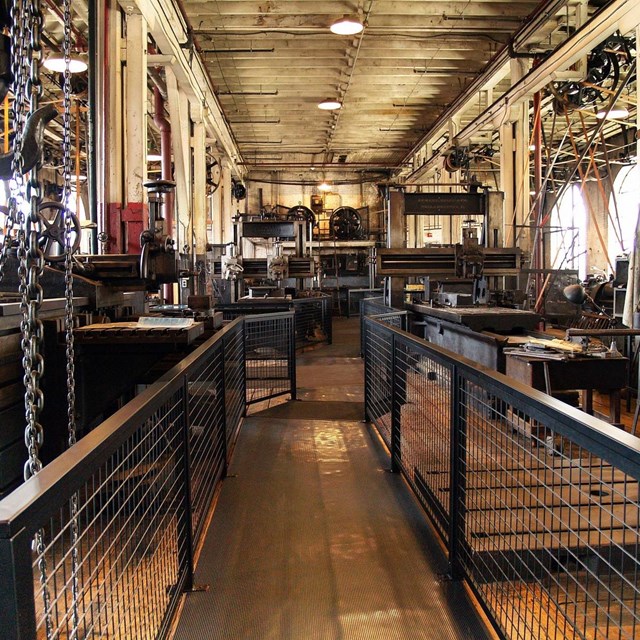 Metal fences guide you through a factory floor where machines driven by belts and chains wait. 