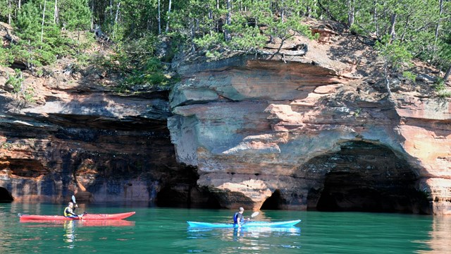 Two kayakers paddle in front of large sandstone cliffs with water carved caves visible.