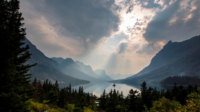 A bolt of sunlight shines through cloudy and hazy skies, hitting mountains and a lake below.