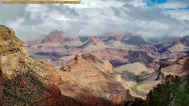 Wispy spring clouds above and partially concealing colorful peaks within a vast canyon landscape.