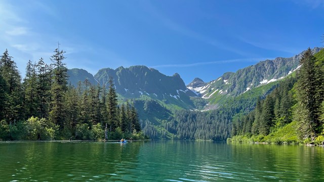 A kayaker paddles in green turquoise waters along forested coast with snow-covered mountains beyond.