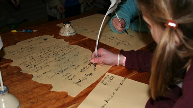 A student writes with a quill pen on parchment in a wooden hut.