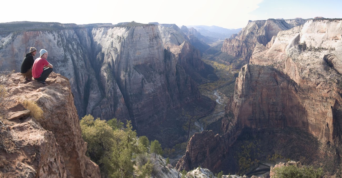 Two people sit on the edge of a deep canyon with a sweeping view from the rim.