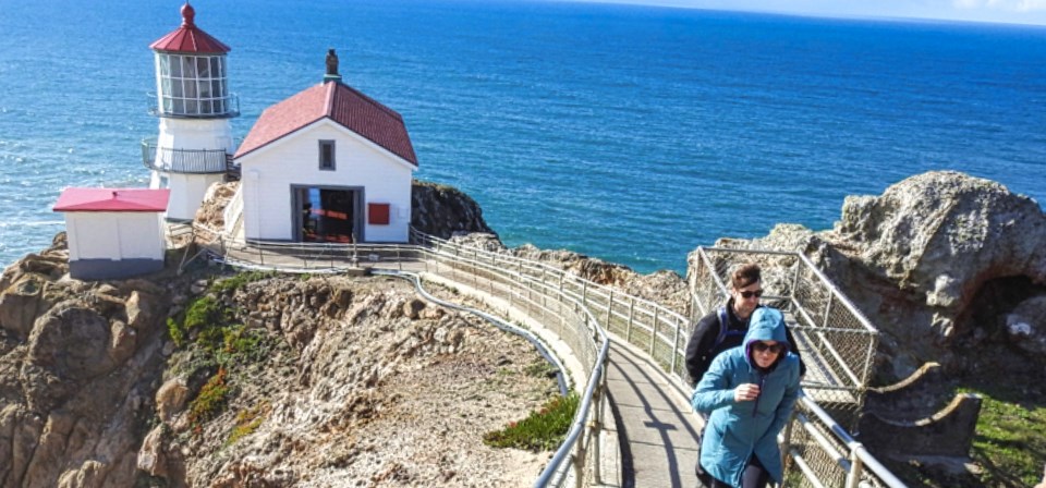 A black and white photo of a wooden ramp running down a rocky, coastal ridge line to a three-story lighthouse tower and two other buildings. A man with three children stand near the base of the stairs.