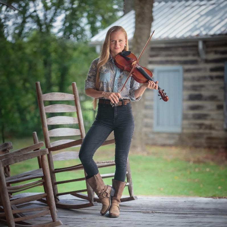 Young lady with blond hair wearing boots plays the fiddle in a rustic setting
