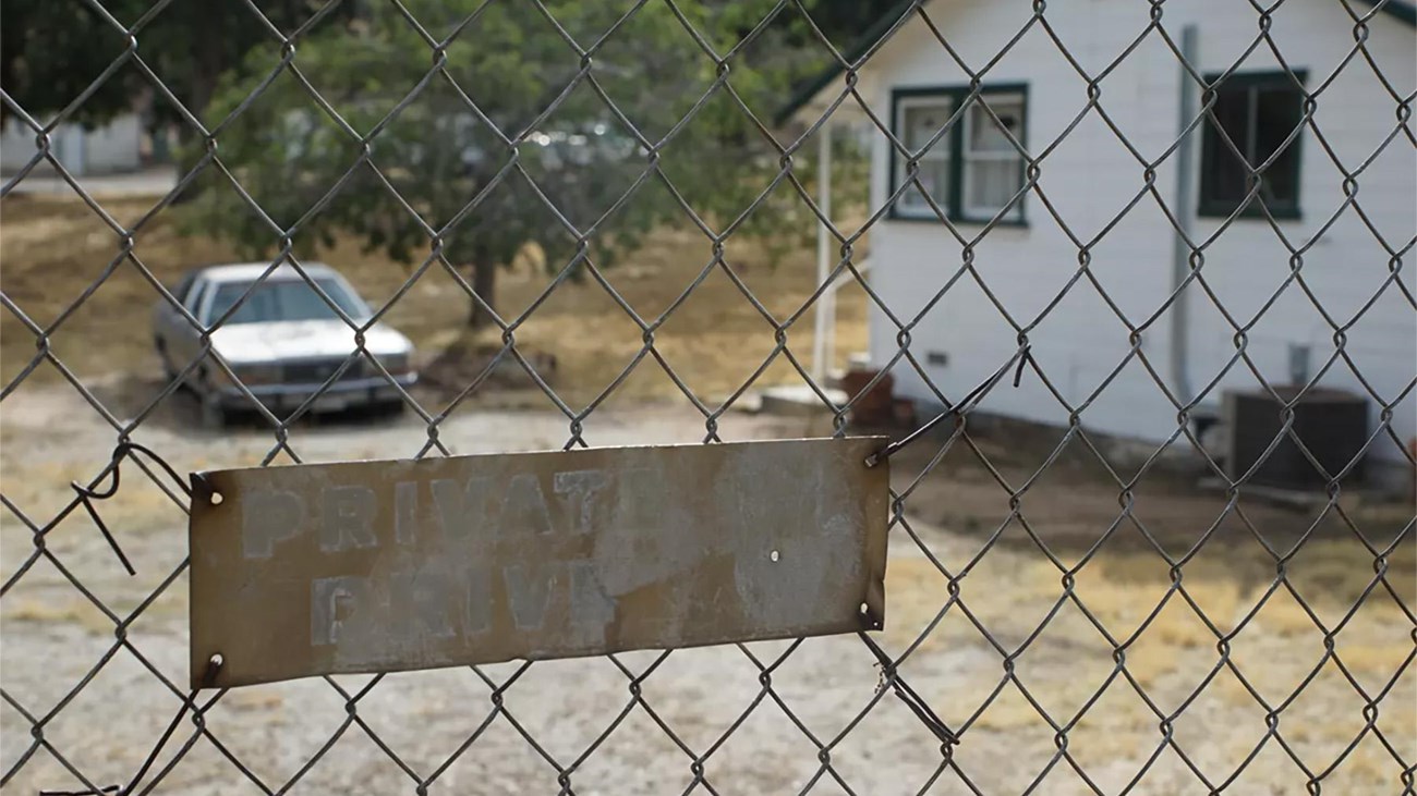 The side of a white house and an old car are background to a close up of a chain link fence section