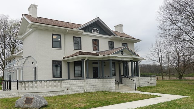 Two story historic brick home pained light gray. Surrounded by green grass. The sky is gray.