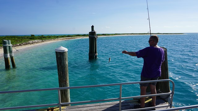 A man standing on a dock by the ocean, holding a fishing pole with a fish on the line.