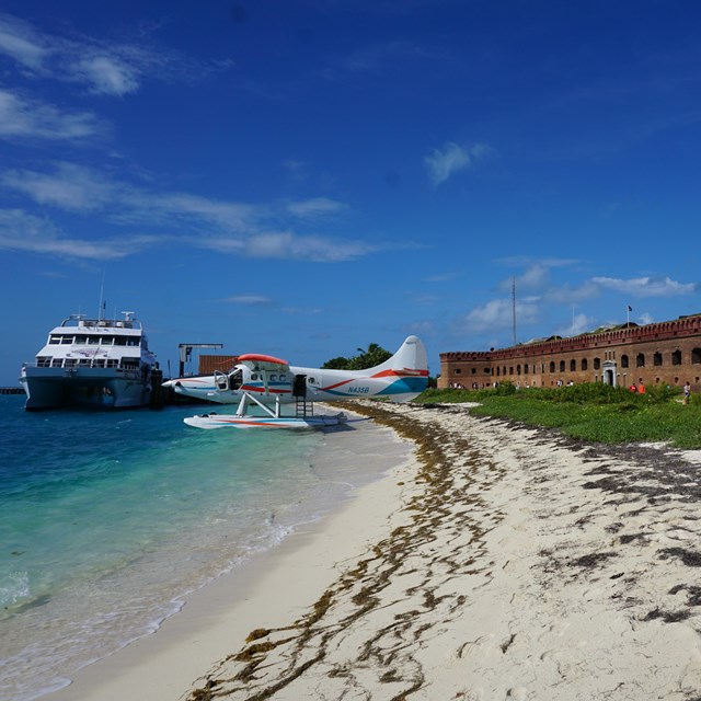 A boat and an airplane docked in front of a beach and a brick structure