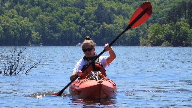 A woman paddling in an orange kayak with rolling hills behind her.