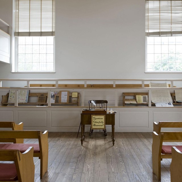 Color photo of rows of wooden pews facing a single desk and chair in the front of the room.