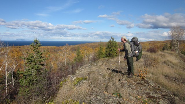 A person with a backpack stands on a rocky ridge looking over a forest and a lake. 