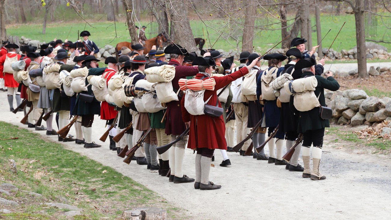 Two lines of 1775 militia soldiers stand shoulder to shoulder placing ramrods into their muskets