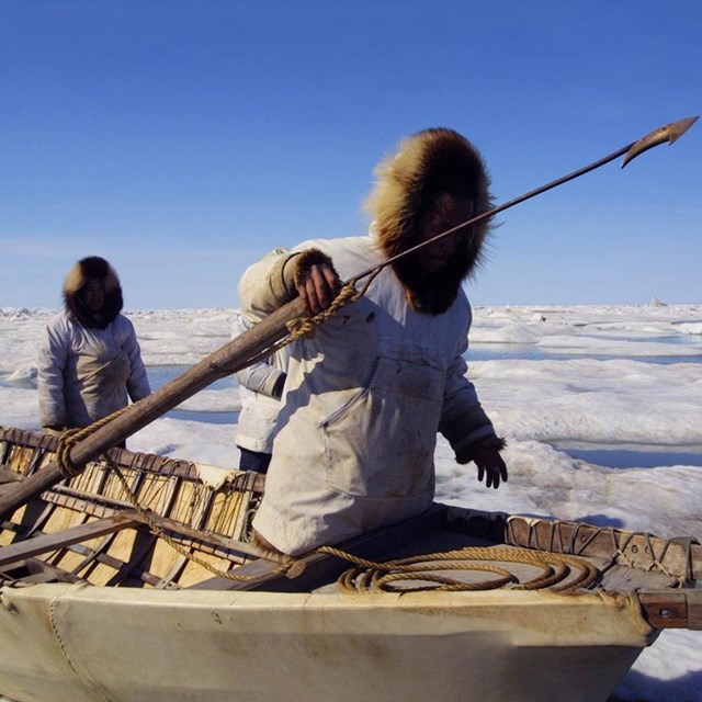 Two whaleman walking along icey paths with a boat and harpoon.