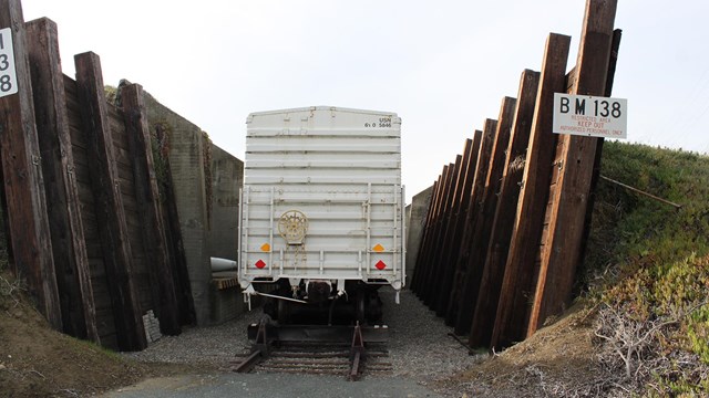 A box car sits on a rail track, between two large wooded walls, backed by earth mounds. 