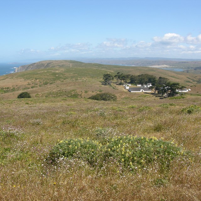 A ranch complex with white wooden buildings on a narrow peninsula between the ocean and a bay.